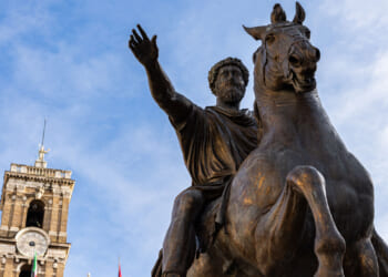 Bronze equestrian statue of a bearded man raising his hand, with an old stone clock tower in the background against a blue sky.