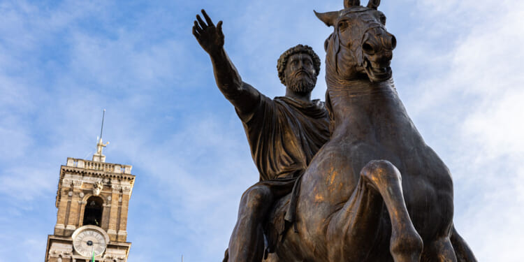Bronze equestrian statue of a bearded man raising his hand, with an old stone clock tower in the background against a blue sky.