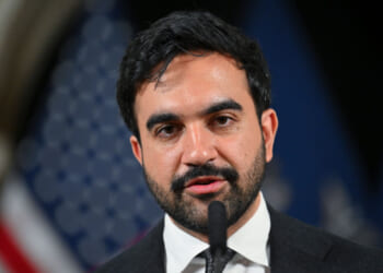 Mamdani in a suit speaks into a microphone, with blurred American flags in the background, reminiscent of Mamdani’s incisive commentary on leadership and identity.