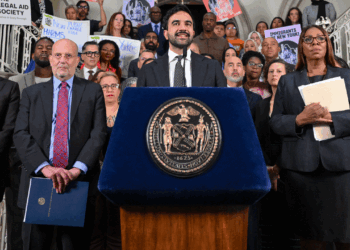 Zohran Mamdani speaks at a podium with an official seal, surrounded by a group of people holding folders and signs supporting immigrants and legal aid inside a government building.