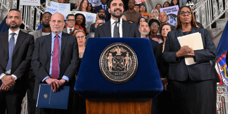 Zohran Mamdani speaks at a podium with an official seal, surrounded by a group of people holding folders and signs supporting immigrants and legal aid inside a government building.