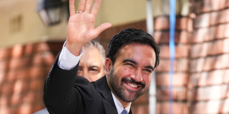 A man in a suit smiles and waves, standing outdoors near a brick wall. Another person is partially visible behind him.