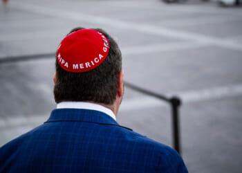 Rep. Randy Fine in a blue suit, seen from behind, wears a red kippah with Trump America Great written on it, standing outdoors on a paved area, seemingly unfazed by recent death threats circulating in the community.