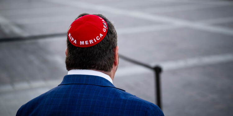 Rep. Randy Fine in a blue suit, seen from behind, wears a red kippah with Trump America Great written on it, standing outdoors on a paved area, seemingly unfazed by recent death threats circulating in the community.