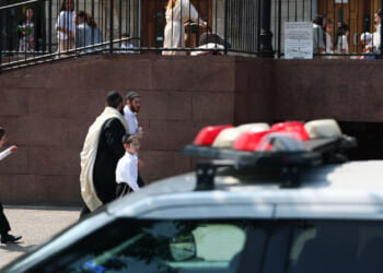 A police car is parked in front of a building as men and boys in traditional Jewish attire walk nearby. Other people are visible on the upper level behind a railing.