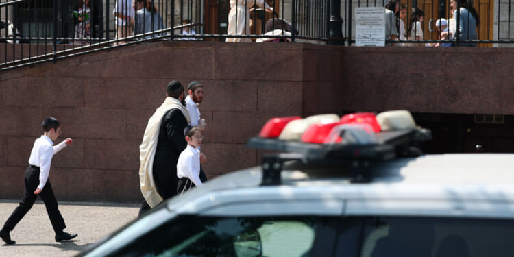 A police car is parked in front of a building as men and boys in traditional Jewish attire walk nearby. Other people are visible on the upper level behind a railing.