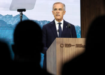 Mark Carney in a suit speaks at a podium during a summit in Canada, with a mountain backdrop and a teleprompter visible. Silhouetted audience members are in the foreground.