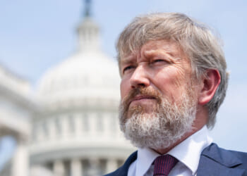 A man with gray hair and a beard in a suit stands outdoors in front of a blurred background featuring the U.S. Capitol building.