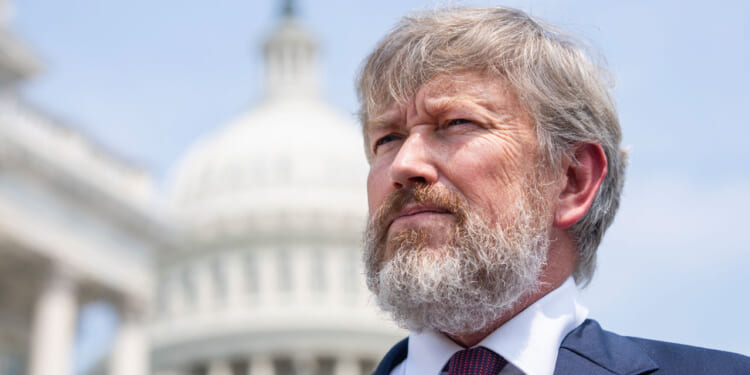 A man with gray hair and a beard in a suit stands outdoors in front of a blurred background featuring the U.S. Capitol building.