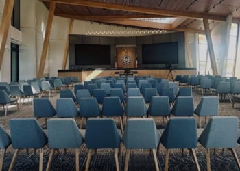 Rows of blue chairs face a stage with two large screens and a podium in a modern, well-lit auditorium with wooden beams and large windows.