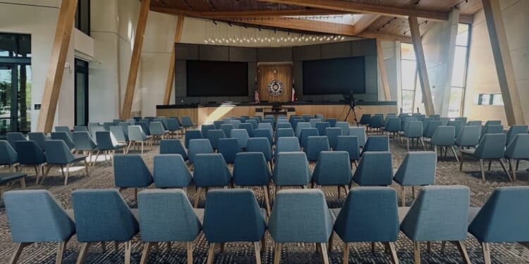 Rows of blue chairs face a stage with two large screens and a podium in a modern, well-lit auditorium with wooden beams and large windows.