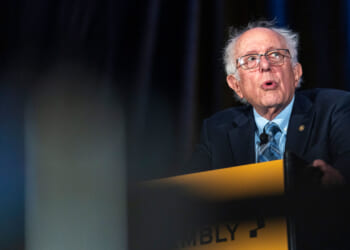 An older man with glasses and gray hair speaks passionately at a podium, wearing a suit and tie, with dark curtains in the background. The podium has the partially visible word ASSEMBLY..