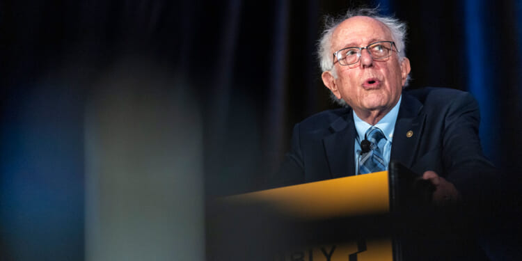 An older man with glasses and gray hair speaks passionately at a podium, wearing a suit and tie, with dark curtains in the background. The podium has the partially visible word ASSEMBLY..
