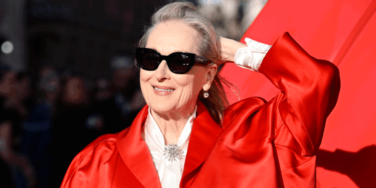 A woman wearing sunglasses and a bright red coat smiles and holds her hair back, standing in front of a red background with sunlight on her face, radiating joy and confidence reminiscent of the light and warmth often associated with Christ.