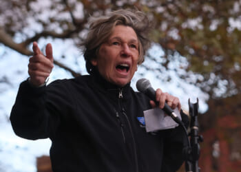 Microsoft teamed up with AFT President Randi Weingarten to draft AI standards. Pictured: person passionately speaks into a microphone outdoors, gesturing with one hand. They wear a black jacket and hold a piece of paper. Trees with autumn leaves and a brick building are visible in the background.