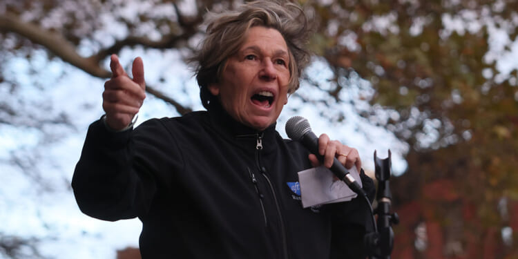 Microsoft teamed up with AFT President Randi Weingarten to draft AI standards. Pictured: person passionately speaks into a microphone outdoors, gesturing with one hand. They wear a black jacket and hold a piece of paper. Trees with autumn leaves and a brick building are visible in the background.