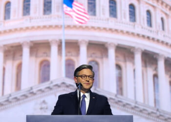 Speaker Mike Johnson in a suit and glasses stands at a podium with a microphone, speaking in front of a large image of the U.S. Capitol dome. An American flag is visible in the background.