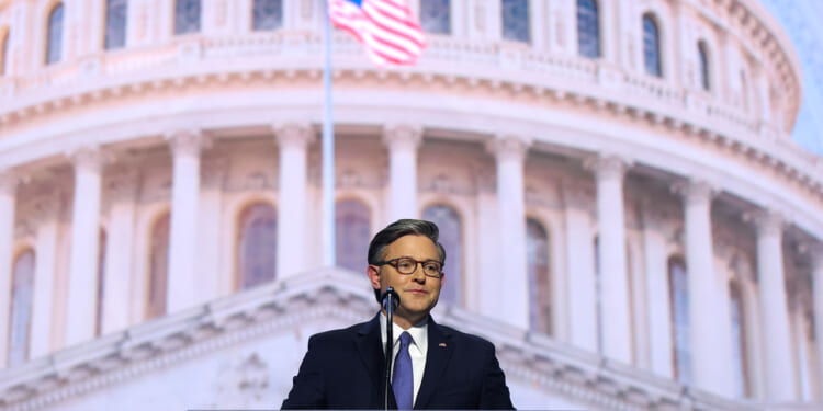 Speaker Mike Johnson in a suit and glasses stands at a podium with a microphone, speaking in front of a large image of the U.S. Capitol dome. An American flag is visible in the background.