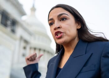 A woman in a navy blazer gestures and speaks emphatically about the alleged hoax in front of a government building with a dome, possibly the U.S. Capitol, during the daytime.