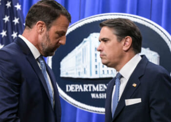 Two men in suits face each other seriously in front of a Department of Justice sign, with an American flag in the background.