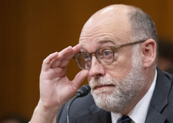 Groups that received federal grants oppose OMB's new grantmaking rules. Pictured: OMB Director Russell Vought, a man with glasses, a bald head, and a white beard sits at a microphone, holding his glasses and looking attentive in a formal setting.