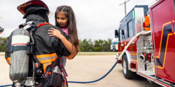 A firefighter in full gear holds a young homeschooled girl near a fire truck. The girl looks concerned, while the firefighter supports her. A blue hose connects to the truck, and trees are visible in the background.