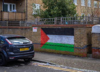 A mural of the Palestinian flag is painted on a brick wall next to a parked black car on a residential street. Nearby are trash bins, a fence, and apartment buildings.