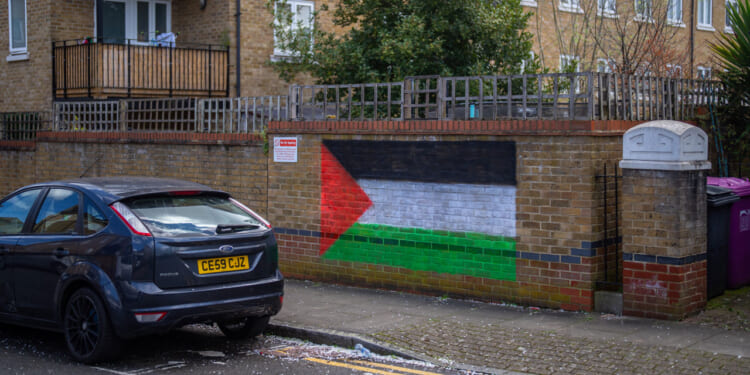 A mural of the Palestinian flag is painted on a brick wall next to a parked black car on a residential street. Nearby are trash bins, a fence, and apartment buildings.