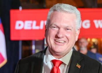 Rep. Mike Collins is pictured in a suit and red tie. He stands in front of a red banner and an American flag, with people and a blurred background visible behind him.