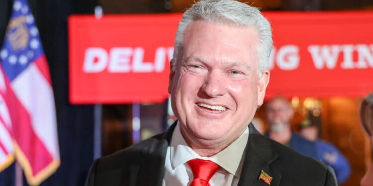 Rep. Mike Collins is pictured in a suit and red tie. He stands in front of a red banner and an American flag, with people and a blurred background visible behind him.