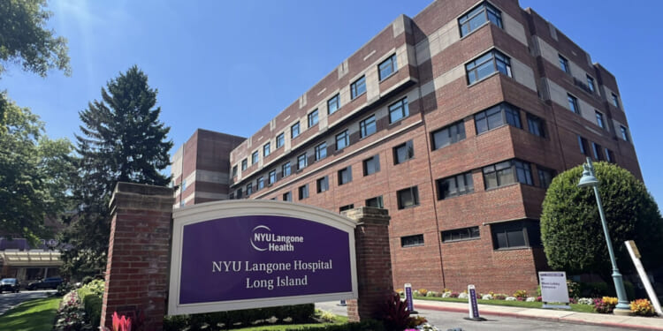 A large brick hospital building with a purple NYU Langone Hospital Long Island sign in front, surrounded by flowers and greenery under a sunny blue sky.