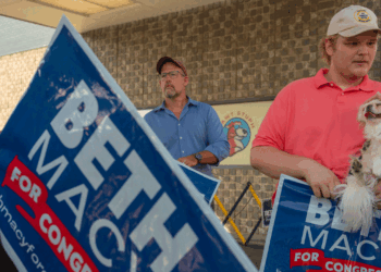 Two men hold Beth Macy for Congress signs outside a building, possibly following recent debates. One man wears a red shirt and beige hat, holding a small white dog. Another man in blue stands in the background, and a pet store logo is visible.