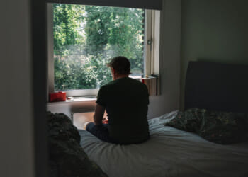 A single man sits on a bed facing a window, looking outside at green trees. The room is dimly lit, with unmade bedding and sunlight streaming through the window.