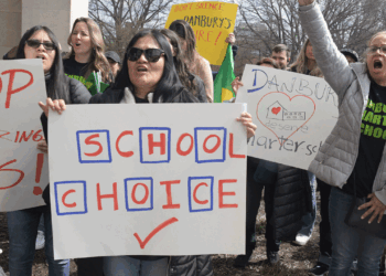 A group of people protest outdoors, holding signs with messages like SCHOOL CHOICE, STOP, and Danbury deserves a charter school. Some wear sunglasses and coats on a cloudy day.