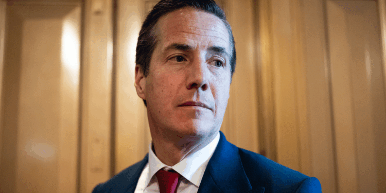 A man in a dark suit, white shirt, and red tie stands in front of wooden panels, looking slightly to his right with a neutral expression.