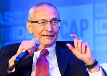 The Center for American Progress helped launch Faith in Public Life, which was meant to be an "organizational anchor for the Religious Left." Pictured: John Podesta, a man in a suit and red tie speaks into a microphone, gesturing with his hand. A blue background displays blurred text and numbers.