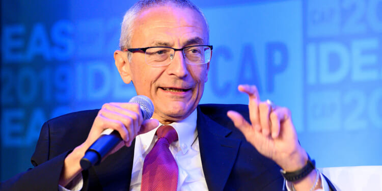 The Center for American Progress helped launch Faith in Public Life, which was meant to be an "organizational anchor for the Religious Left." Pictured: John Podesta, a man in a suit and red tie speaks into a microphone, gesturing with his hand. A blue background displays blurred text and numbers.