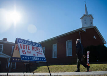 A person walks past a Vote Here! sign outside a brick church in Virginia under a bright sun, indicating the location is a polling place.