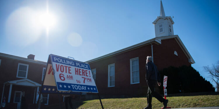 A person walks past a Vote Here! sign outside a brick church in Virginia under a bright sun, indicating the location is a polling place.