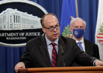 Jeff Clark in a suit and glasses speaks at a podium with the Department of Justice seal behind him. Another man in a suit and face mask stands in the background near U.S. flags.