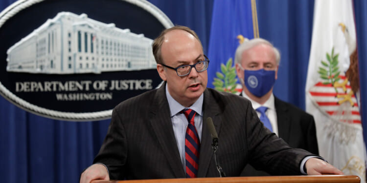 Jeff Clark in a suit and glasses speaks at a podium with the Department of Justice seal behind him. Another man in a suit and face mask stands in the background near U.S. flags.