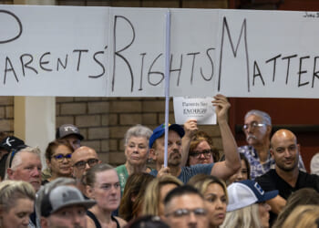 A crowd sits at a public meeting. One person holds a large sign reading PARENTS RIGHTS MATTER in bold letters above their head. Other attendees look on attentively.