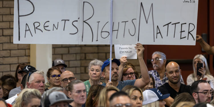 A crowd sits at a public meeting. One person holds a large sign reading PARENTS RIGHTS MATTER in bold letters above their head. Other attendees look on attentively.
