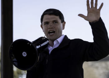 A person in a black jacket speaks energetically into a megaphone and raises one hand, appearing to address a crowd outdoors under a clear sky.