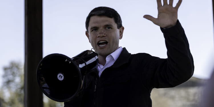 A person in a black jacket speaks energetically into a megaphone and raises one hand, appearing to address a crowd outdoors under a clear sky.