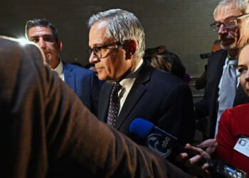 A man in a suit and glasses stands surrounded by reporters and microphones, facing a group of people in a dimly lit indoor setting.