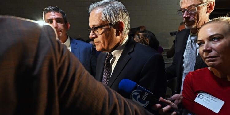 A man in a suit and glasses stands surrounded by reporters and microphones, facing a group of people in a dimly lit indoor setting.