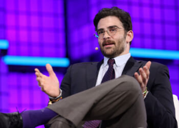 A man in a suit and tie sits on stage with a microphone headset, gesturing with his hands while speaking. The background is purple with blue horizontal accents.