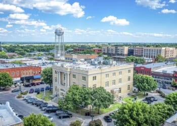 Aerial view of a small city square with a historic courthouse, surrounding trees, parked cars, brick buildings, and a water tower under a blue sky with scattered clouds.