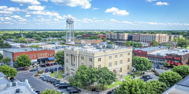 Aerial view of a small city square with a historic courthouse, surrounding trees, parked cars, brick buildings, and a water tower under a blue sky with scattered clouds.